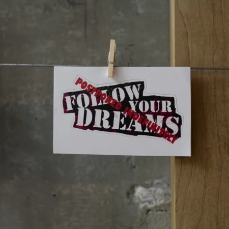 Sticker that reads FOLLOW YOUR DREAMS With Postponed indefinitely stamped overtop in red.